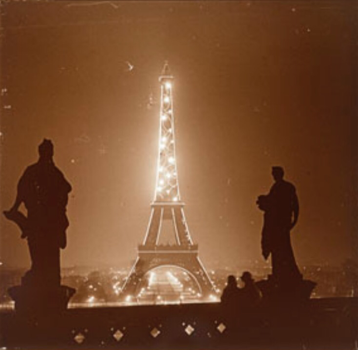 La Tour Eiffel illuminée la nuit, avec deux statues en premier plan, vers 1925